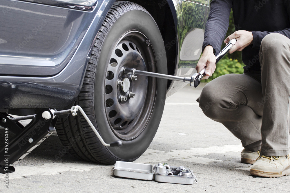 Person changing a flat tire on the side of the road, using a jack and lug wrench.